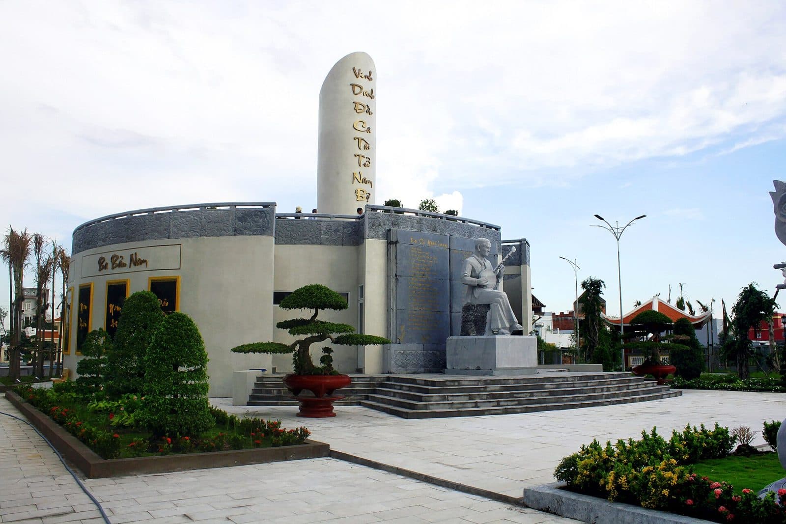 Tượng đá nhạc sĩ Cao Văn Lầu ngồi ôm đờn kìm, phía sau là khối nhà tròn và trụ đá khắc dòng chữ vinh danh đờn ca tài tử Nam Bộ